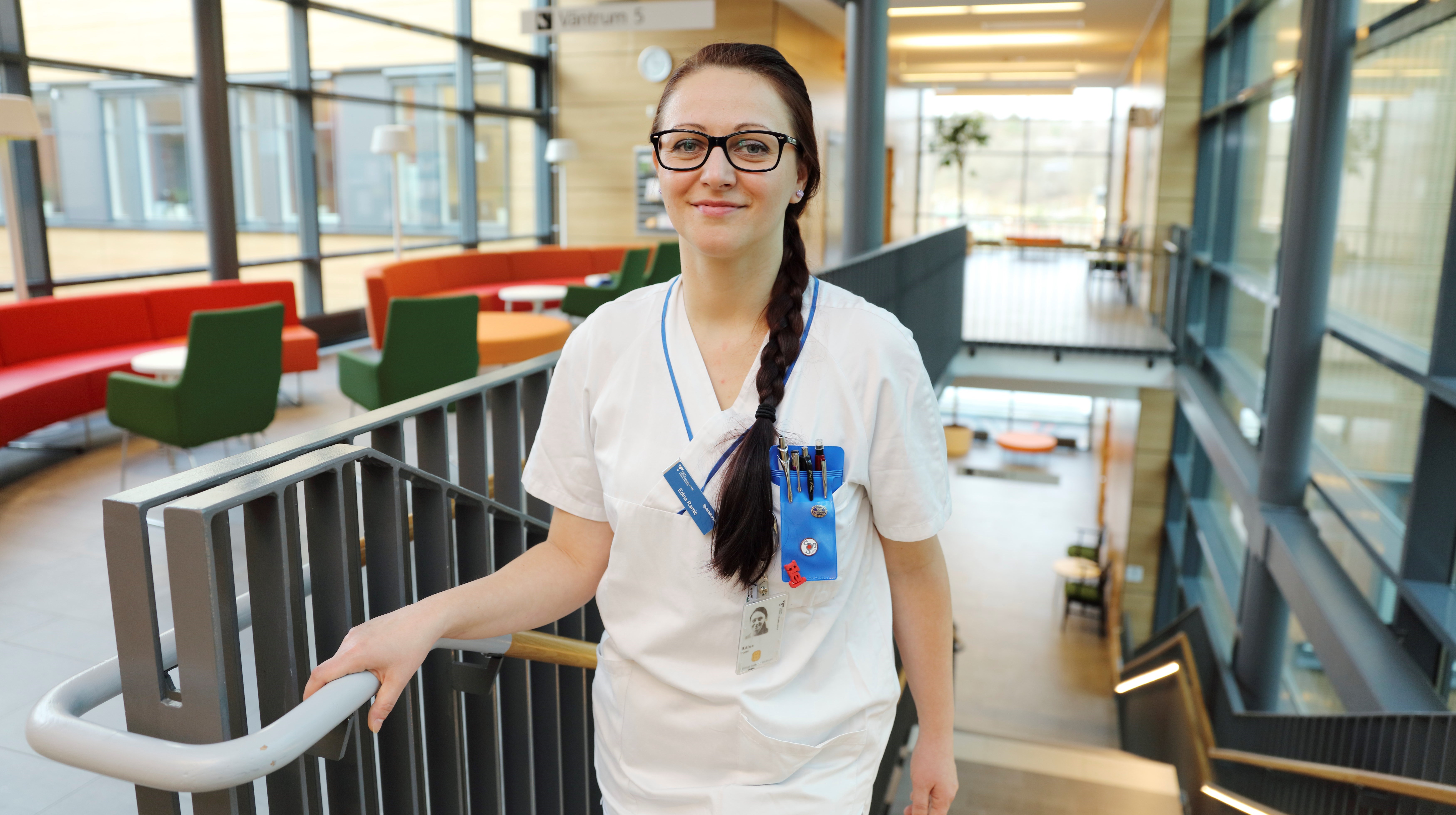 En sjuksköterska med lång mörk fläta och glasögon står längst upp i en trappa i ett sjukhus 
