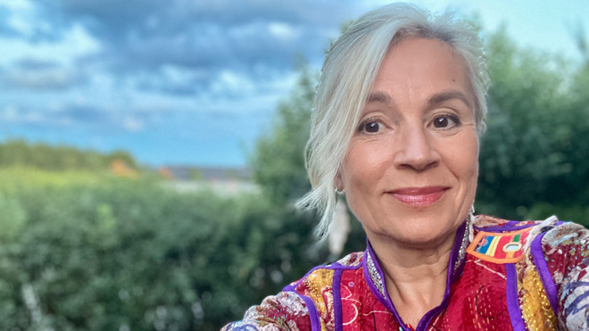 Susanne Redebo är i medelåldern. Hon har långt grått hår som är uppsatt och kammat i sidbena. Hennes ögon är bruna och hon är klädd i en färgglad blus. Hon står utomhus, bakgrunden är suddig med grönska och en blå himmel med moln. Hon ler med stängd mun.