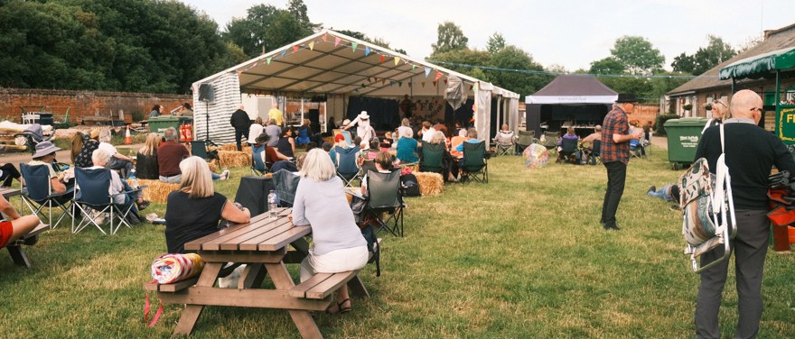 Utspridd publik med ryggarna mot kameran riktar uppmärksamheten till ett stort vitt evenemangstält. Utomhus på gräsmatta, publiken sitter på campingstolar och höbalar.