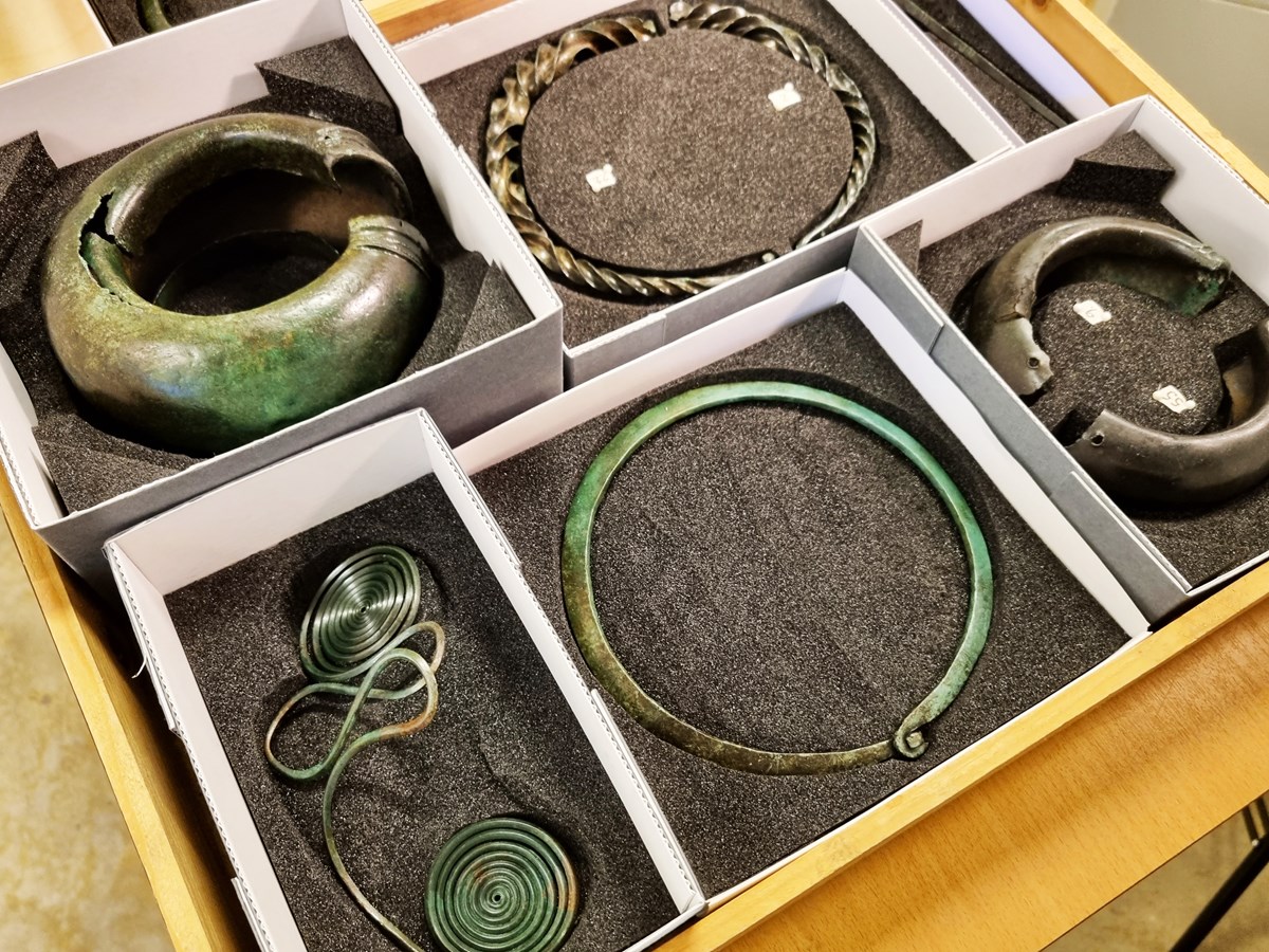 Fem olika föremål i brons ligger i ett skyddande material i varsin låda. Föremålen är skimrade gröna och består bland annat av två olika halsband. 