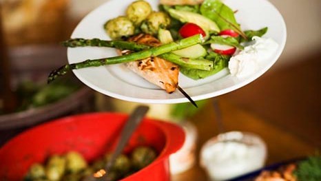 En tallrik med en portion upplagd av lax och grönsaker. På bordet syns en röd bunke med potatis och en plåt med lax.