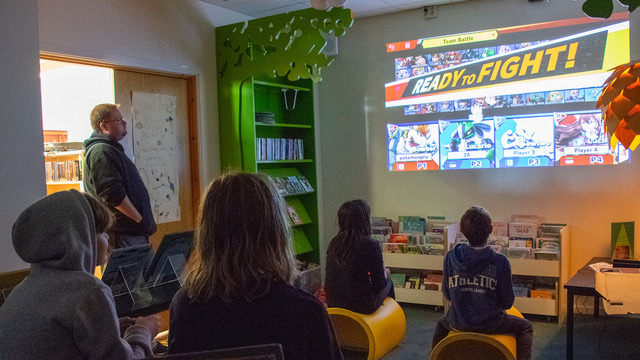 Fyra barn sitter i ett nedsläckt rum. På en vägg finns ett spel projicerat, det står "Ready to fight!" på väggen. Barnens ögon är fästa på väggen, de har spelkontroller i sina händer. Biblioteksassistenten Emil Andersson står i dörröppningen och kollar på spelet. Runt om i rummet finns bokhyllor och boktråg med barnböcker.