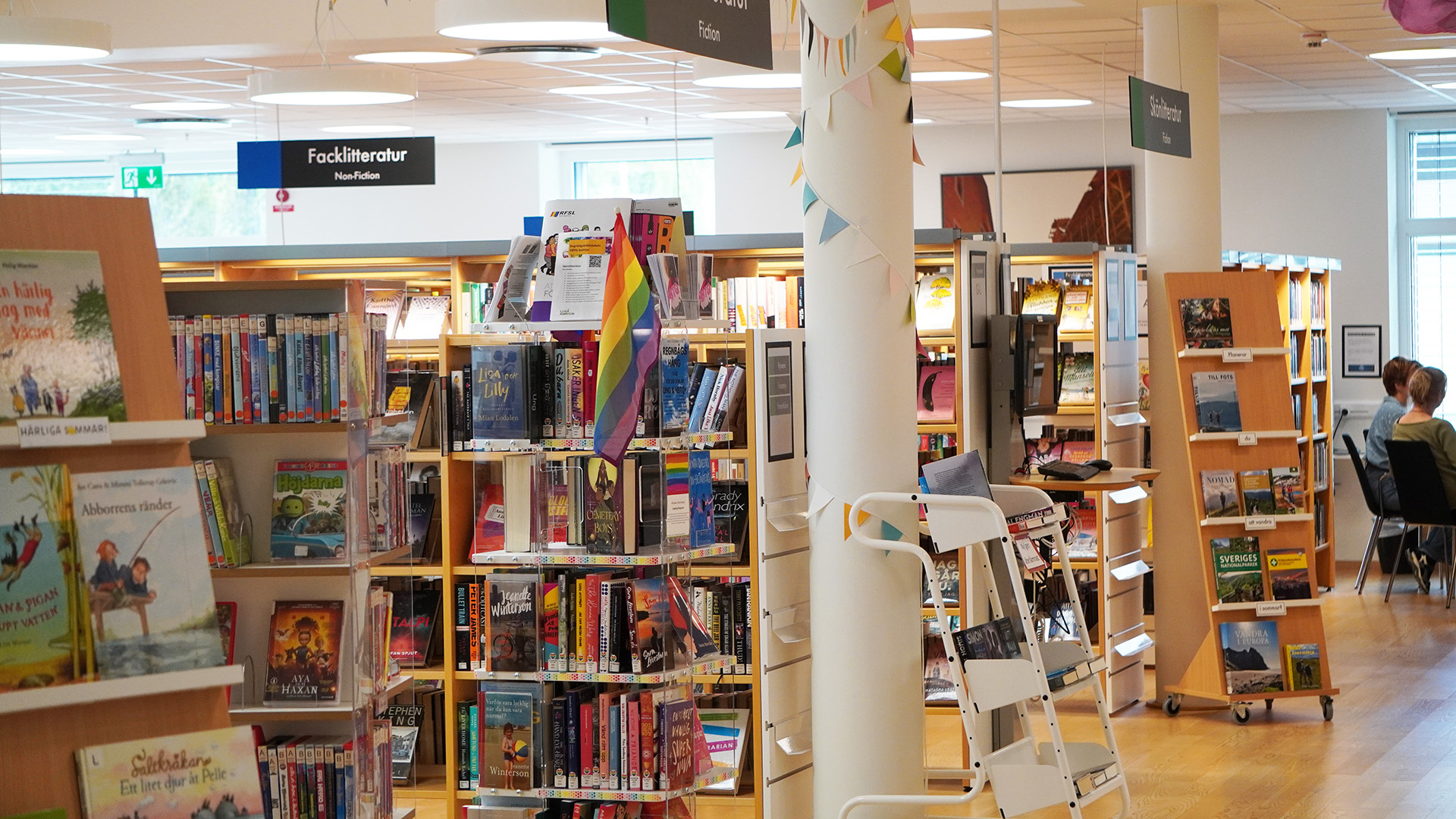 Vännäs bibliotek. Bokhyllor i ljust trä står på rad, på en skylt i taket står det Facklitteratur. Vita pelare står utmed en gång, vid ett fönster längst bort sitter två personer och samtalar.