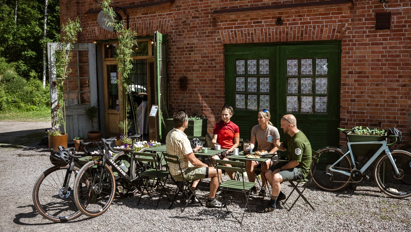 En grupp på fyra personer sitter i solkskenet utanför en röd tegelbyggnad och samtalar vid ett cafébord