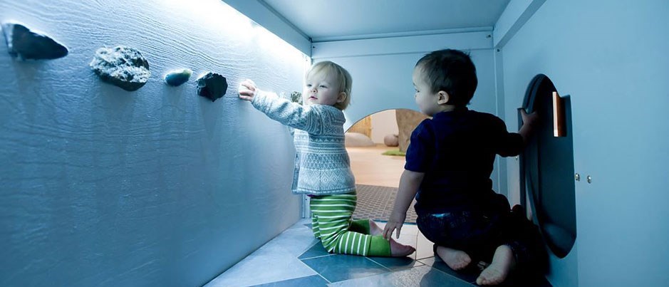 Två små barn sitter i en liten tunnel och leker med stenar som sitter på väggen.