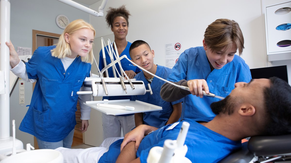 Tre elever, en flicka och två pojkar iklädda tandvårdens blå arbetskläder besöker en kvinnlig tandläkare och en manlig tandhygienist. En av eleverna testar ett redskap på tandhygienisten när han ligger i tandläkarstolen.