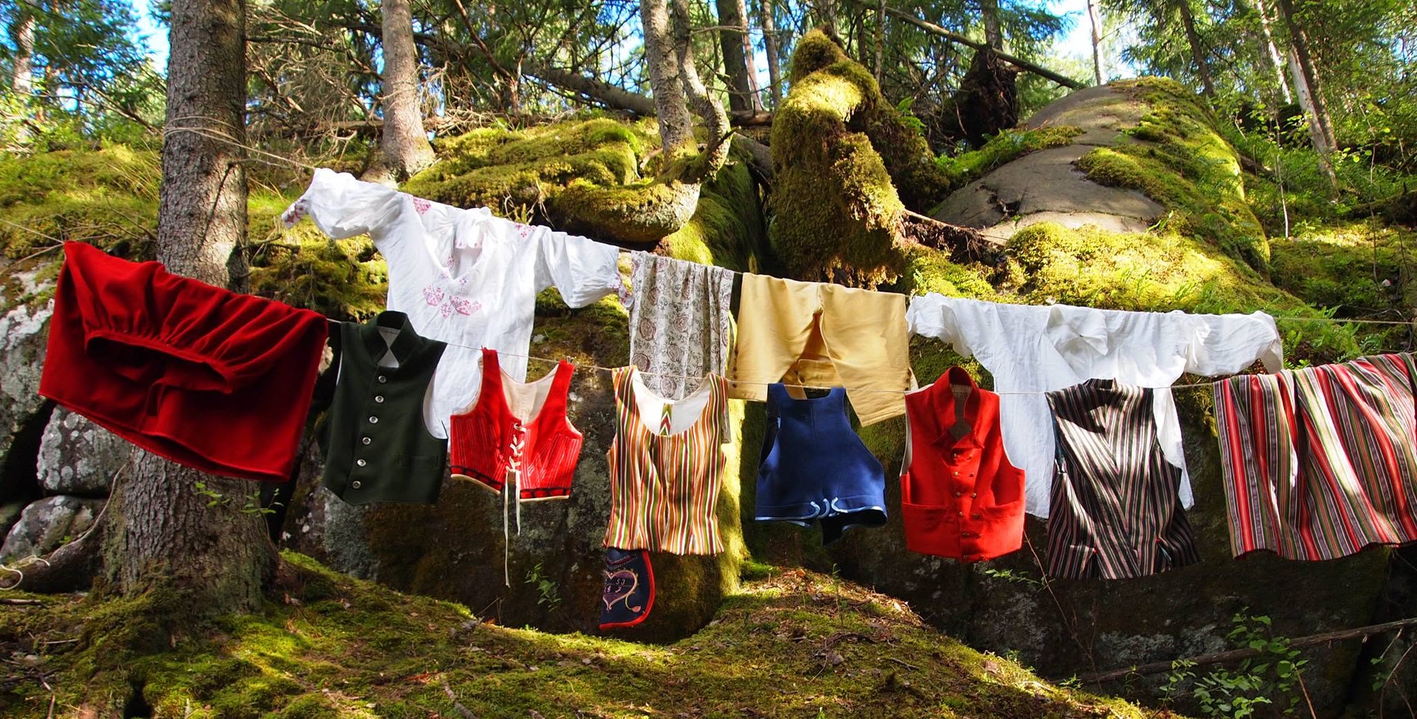 Folkdräktsdelar som hänger på tvättlinor ute i skogen med mossiga stenar och stora träd i bakgrunden: Vita överdelar med broderier, förkläde med tryckt blommönster, mansbyxor i gult, yllekjolar en röd och en randig, mansvästar en grön, en röd och en randig, kvinnolivstycken ett rött, ett blått och ett randigt. 